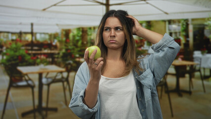 Fototapeta premium Woman holding green apple with hand to head on street terrace wearing denim shirt and white tanktop; uncertainty decision.