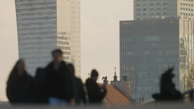 Urban lifestyle scene with blurred foreground pedestrians in motion, sidewalk activity and commuters on their way, distant warm color glass skyscrapers skyline in crisp focus under soft daylight