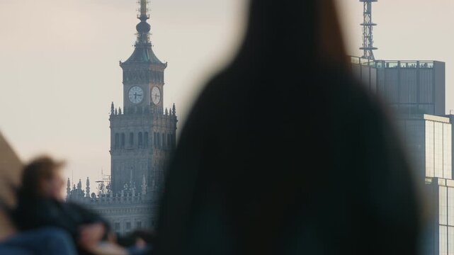 Urban lifestyle scene defocused foreground silhouettes on pedestrian bridge, people cycling and walking, distant skyline of glass skyscrapers and clock tower in focus, warm evening light, spring city