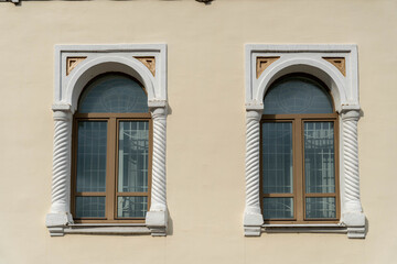 Two Arched Windows On Pale Facade, Ornate Twisted Pilasters And Decorative Cornices Framing Wooden Frames And Reflective Glass, Warm Sunlight Casting © Egor Lint
