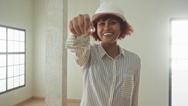 Woman holding keys with outstretched hand in an empty building room, smiling while wearing hardhat and striped shirt as architect presenting access; homeowner pride.
