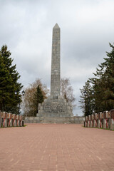 Obraz premium Tall Obelisk Rising Above Memorial Plaza, Photographer Frames Vertical Silhouette, Granite Steps And Sculptural Relief At Base, Pine Rows Flank