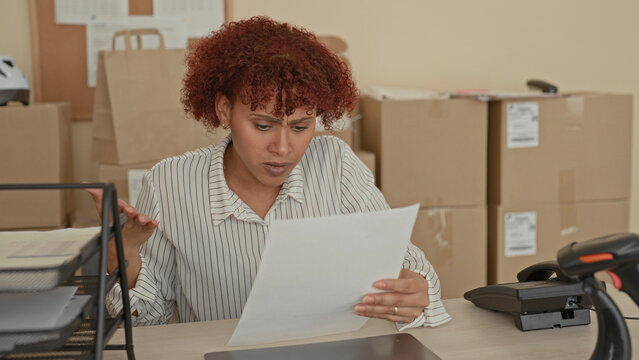 Woman holding document, finger to temple at desk inside building filled with cardboard boxes; small business stress.