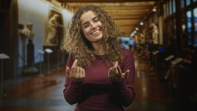 Woman, young with curly hair, beckons with fingers toward marble sculptures in a museum building corridor while smiling and facing camera; playful invitation.