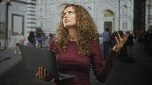 Young woman with curly hair holding laptop, scrolling and gesturing with one hand in front of historic cathedral building; travel work frustration.