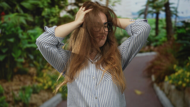Woman with glasses covers ears and holds head amid lush green forest pathway under sunlight; stress.