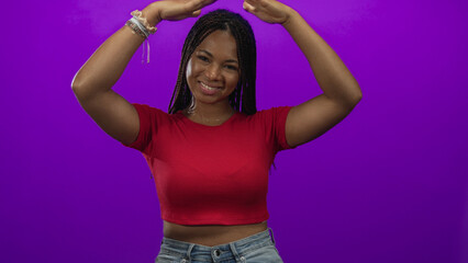 Woman with hands raised above head, bare midriff visible and bracelets, smiling in studio with purple backdrop; joy confidence.