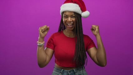 Fototapeta premium Woman making peace sign and cheering with raised fists in studio wearing santa hat and red crop top; holiday joy.