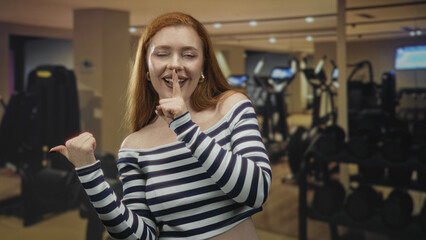 Redhead woman with finger to lips for silence and thumb pointing at dumbbell rack in gym  playful secrecy. © Krakenimages.com