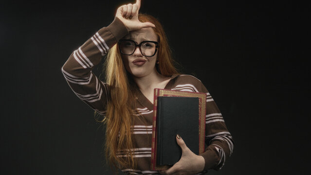 Redhead woman wearing glasses holding book makes l sign on forehead then points finger to camera in studio; mockery accusation.