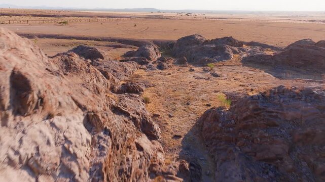 FPV drone navigates through weathered brown rock formations in vast arid desert plains during golden hour, revealing textured boulders, sparse vegetation, and endless flat terrain under hazy skies.