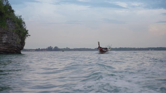 Traditional Thai longtail boat cruises through calm Andaman Sea waters past dramatic limestone karst cliff covered in lush green vegetation under soft overcast evening sky in Krabi province.