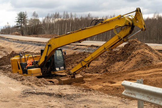 Excavator Digging Trench Beside Road, Yellow Excavator Arm Scooping Sandy Soil, Deep Rut With Piled Spoil, Overcast Sky, Bare Birch Trees