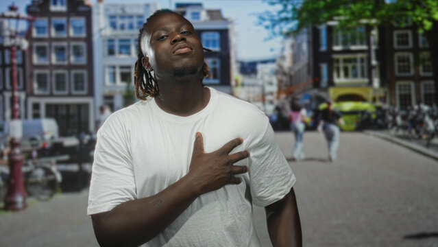 Young african american man clutching chest and bending forward on a busy city street with bicycles and canal bridge visible; pain emergency.