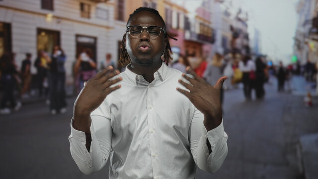 Man gesturing with hands and touching temple on a busy city street, wearing white shirt and glasses; passionate speech.