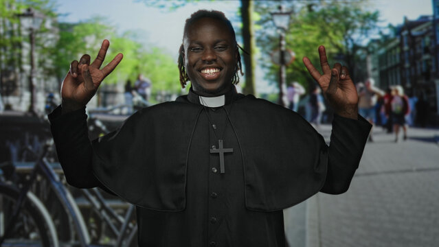 Man priest in cassock holds up peace sign with both hands on a busy street; joy faith community hope.