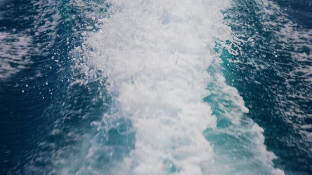 Churning white foam and spray create a dramatic wake trail behind a fast moving boat on deep turquoise ocean water. Bubbles and splashes form dynamic patterns on the surface.