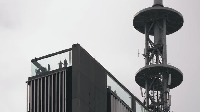 Low angle view of skyscraper rooftop observation deck with people behind glass, city viewing platform activity, tower top against cloudy sky and open space, modern architecture concept
