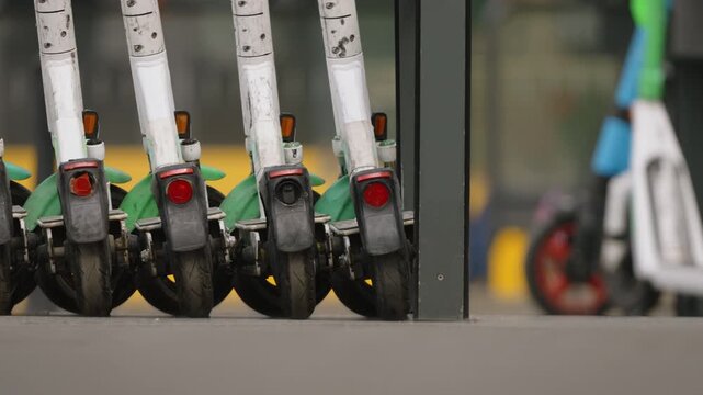 Ground level close up of rental electric scooters parked in a row at charging station, urban street activity in background with cars and pedestrians, modern city mobility concept