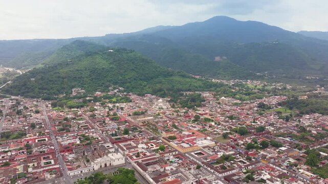 Drone footage captures stunning aerial panorama of historic colonial town in Guatemala nestled in lush green mountain valley with red rooftops and church buildings visible below cloudy sky.