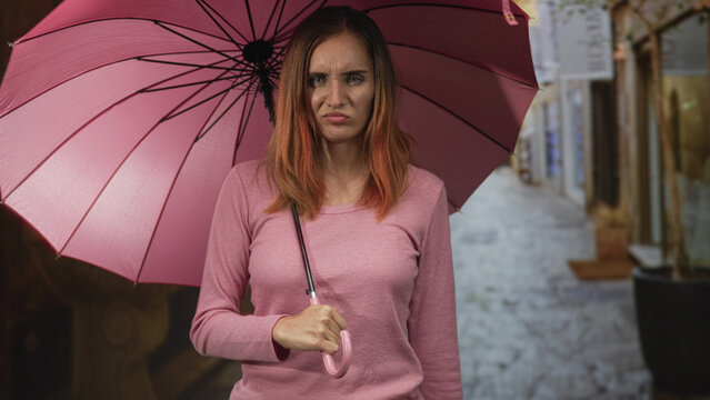 Redhead woman wearing pink sweater, holding pink umbrella and gripping curved handle, grimacing with pursed lips on a narrow cobblestone street between shopfronts; annoyance.