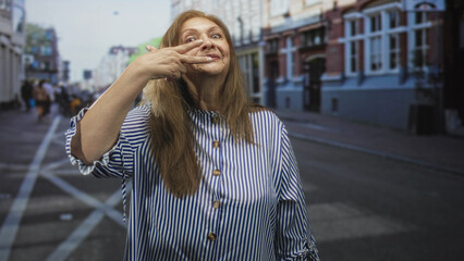 Fototapeta premium Woman senior hispanic shows two fingers over eye with hand visible on urban street, striped blouse and slight smile; playful confidence.