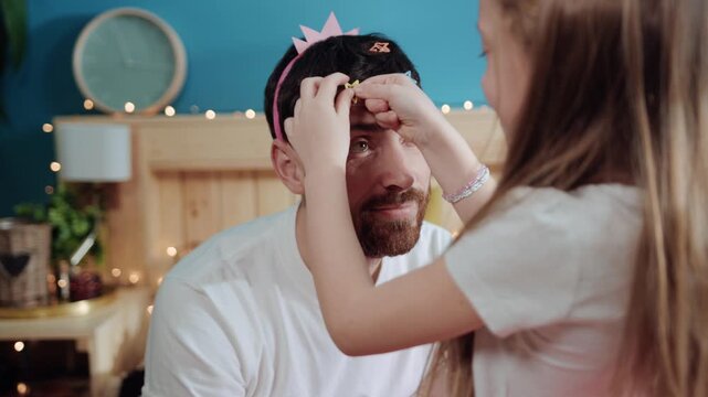 Closeup little daughter princess putting hairpins stars on her father's head for costume party. Happy young daddy waiting dressing with his little child for birthday celebration.