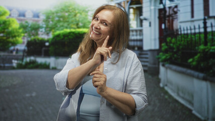 Fototapeta premium Woman smiling with hands pressed together and open palms in alternating gestures wearing light blue top and checked shirt on a residential street; gratitude joy.