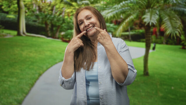 Woman points fingers to mouth, pulling cheeks to show teeth on a park path street lined with palm trees and green lawn; joy warmth connection.