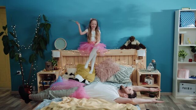 Happy affectionate young family playing together at costume party. Cheerful handsome daddy dressed like princess doing angel moves lying on bed. Cute girl little fairy laughing and smiling.