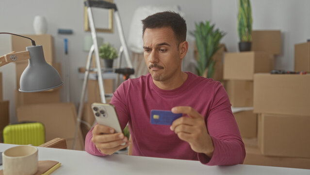 Young man with moustache holds smartphone and creditcard, frowning while checking a charge amid moving boxes in a building; financial frustration.
