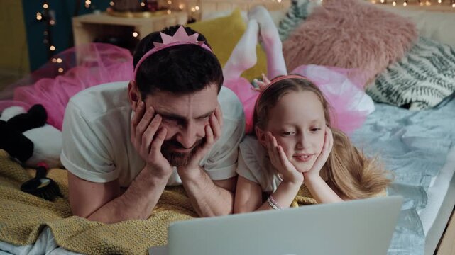 Charming beautiful fairies dad and daughter watching exciting cartoons on laptop resting on bed having a custom disney party at home.