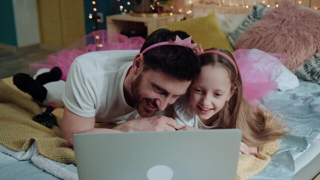Cheerful father and his girl daughter dressed likfe funny pink fairies lying down on bed watching cartoons together laughing and smiling indoors. Happy birthday party.