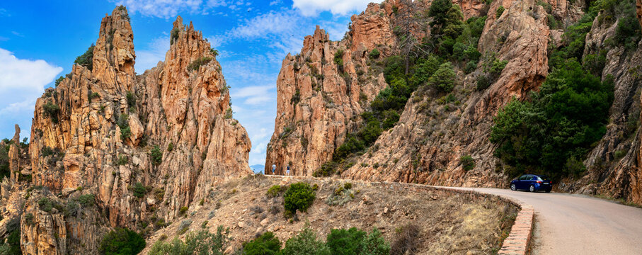 Calanques de Piana Corsica 11k panorama of red granite rocks on D81 road. Scenic mountain pass view of winding highway and car between huge cliffs in France. Mediterranean road trip travel scenery