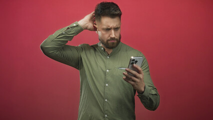 Fototapeta premium Man holding smartphone and touching ear while frowning and scratching his head, checking the screen in a red studio backdrop; confusion.