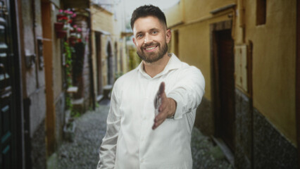 Obraz premium Hispanic man with beard extends hand for handshake in a narrow cobblestone street, wearing white shirt and smiling; friendly trust.