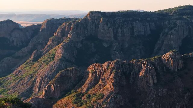 Rugged Sierra Madre Occidental mountain range bathed in warm golden sunset light in Durango, Mexico. Dramatic rocky cliffs with green vegetation create stunning layers of depth across vast landscape.