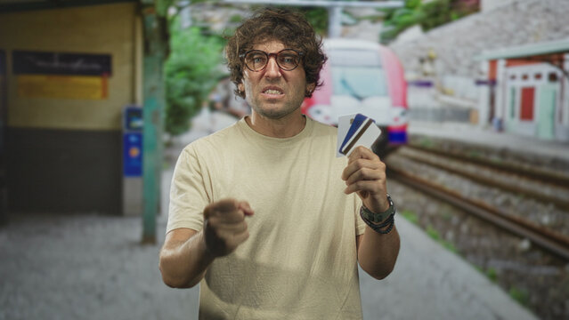 Man grimaces holding creditcard on a train platform inside a building, wearing beige tshirt and glasses and displaying a tense travel gesture; frustration travel delay.