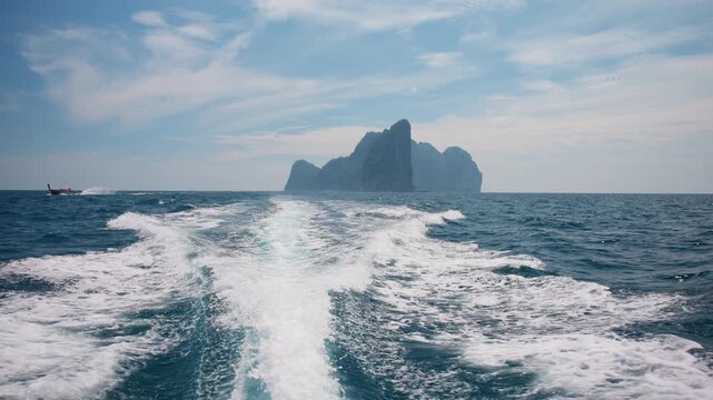 Speedboat wake churns through deep blue Andaman Sea waters as towering limestone cliffs of Phi Phi Islands emerge through tropical haze under wispy clouds and bright afternoon sunlight in Thailand.