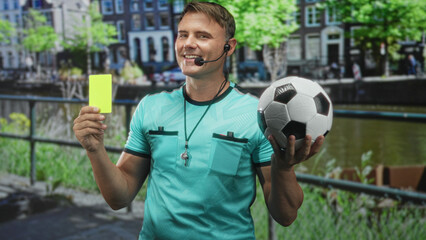 Naklejka premium Man referee holding yellow card and soccer ball with whistle and headset by canal railing on street; confidence fairness.