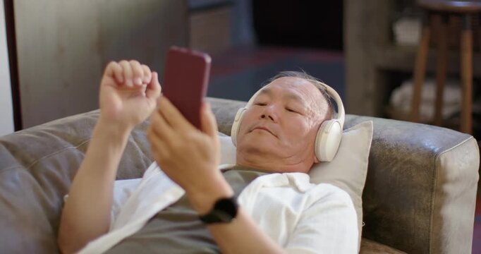 Senior Asian man scrolling phone reacting to alert lowering it closing eyes wearing headset on sofa