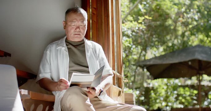 Senior Asian man sitting by open wooden window reading booklet pausing sun drawing gaze to garden