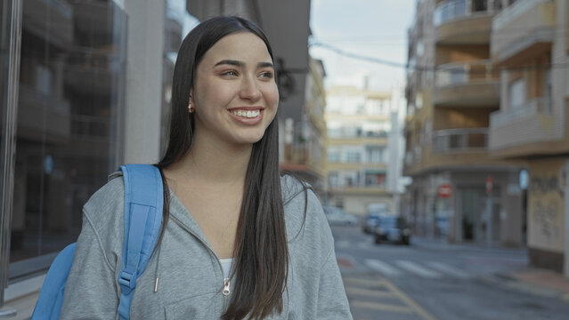 Young hispanic woman wearing a blue backpack smiles while looking to the right on a city street with apartment buildings; optimism.