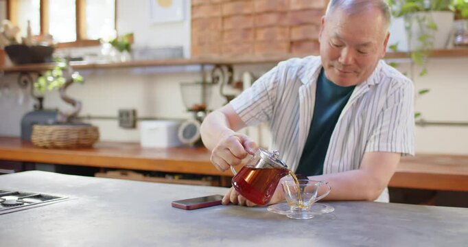 Asian senior man lifting teapot, pouring tea at kitchen counter near smartphone to fill cup