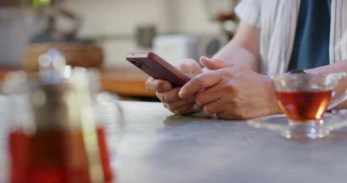 Senior Asian man sitting at counter, picking up smartphone, tapping and reading texts, teacup right