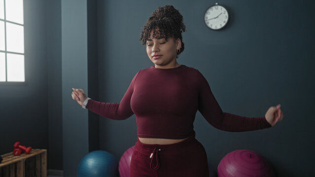 Woman holds shoulder and stretches arm across chest in studio with exercise balls, dumbbells, clock and window light; calm recovery.