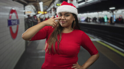Fototapeta premium Woman wearing santa hat points finger to nose on a subway platform inside a building; holiday playful cheer.