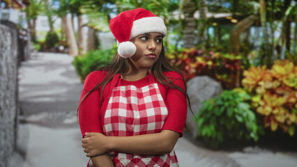 Fototapeta premium Woman wearing santa hat and red gingham apron, arms crossed holding elbow while gazing sideways in studio holiday set; playful holiday mood.