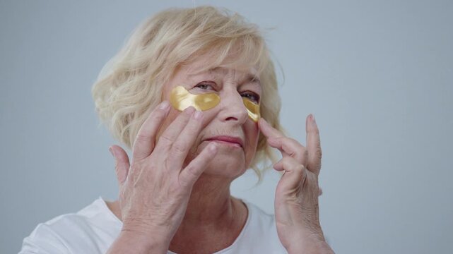 Close-up of beautiful older blonde woman applying eye patches with hands looking in the mirror standing over grey background.
