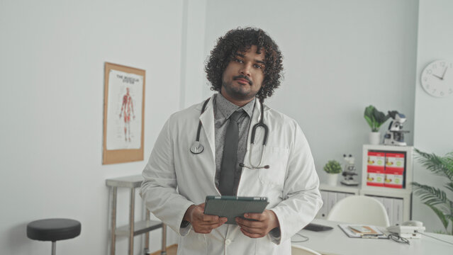 Man doctor in white coat holding a tablet and stethoscope, tapping screen while standing in a clinic building by a desk; calm care.